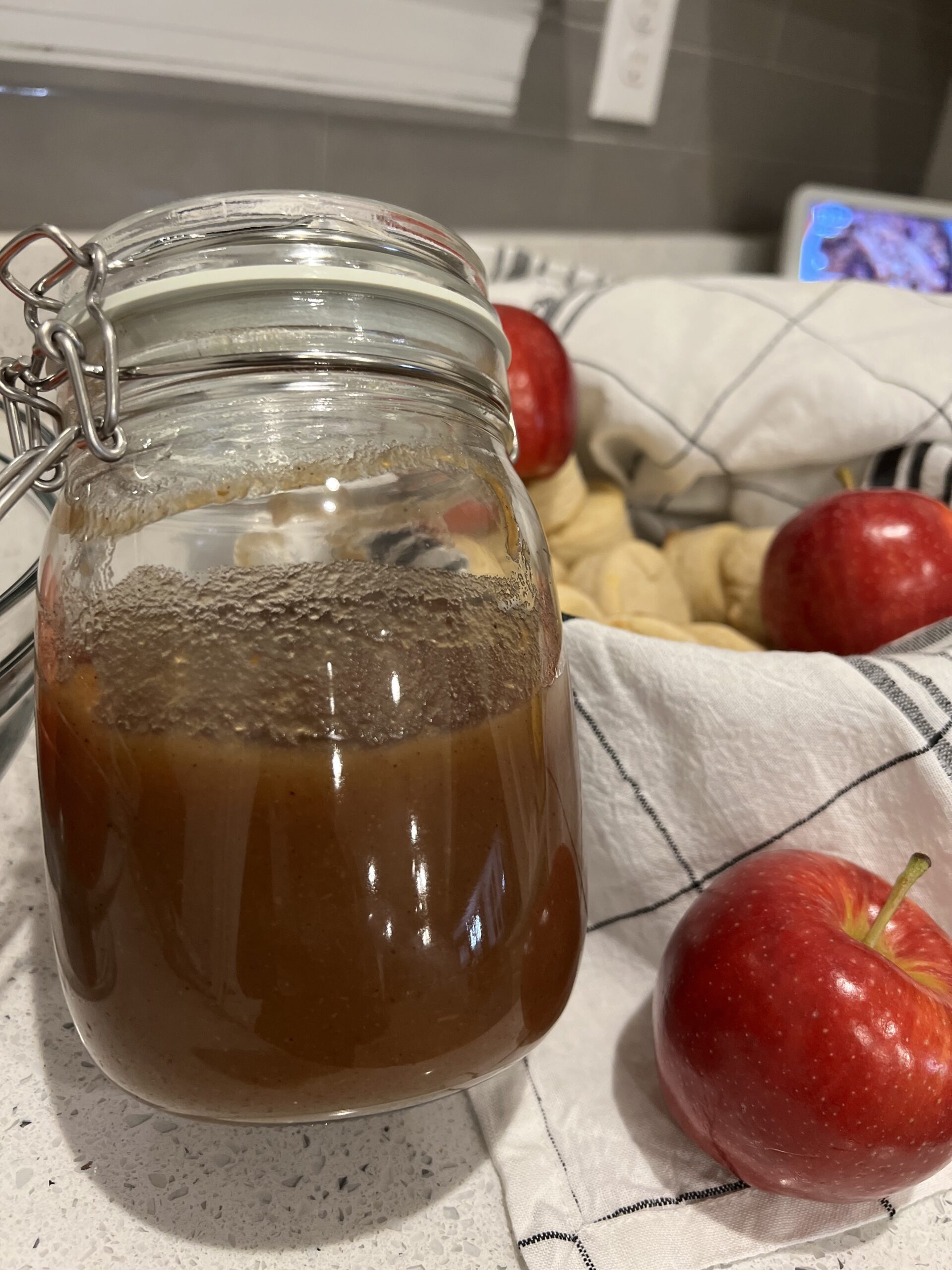 Apple butter inside of a glass mason jar sitting next to a basket of apples and fresh cooked biscuits. The basket is lined with a white and black plaid towel.