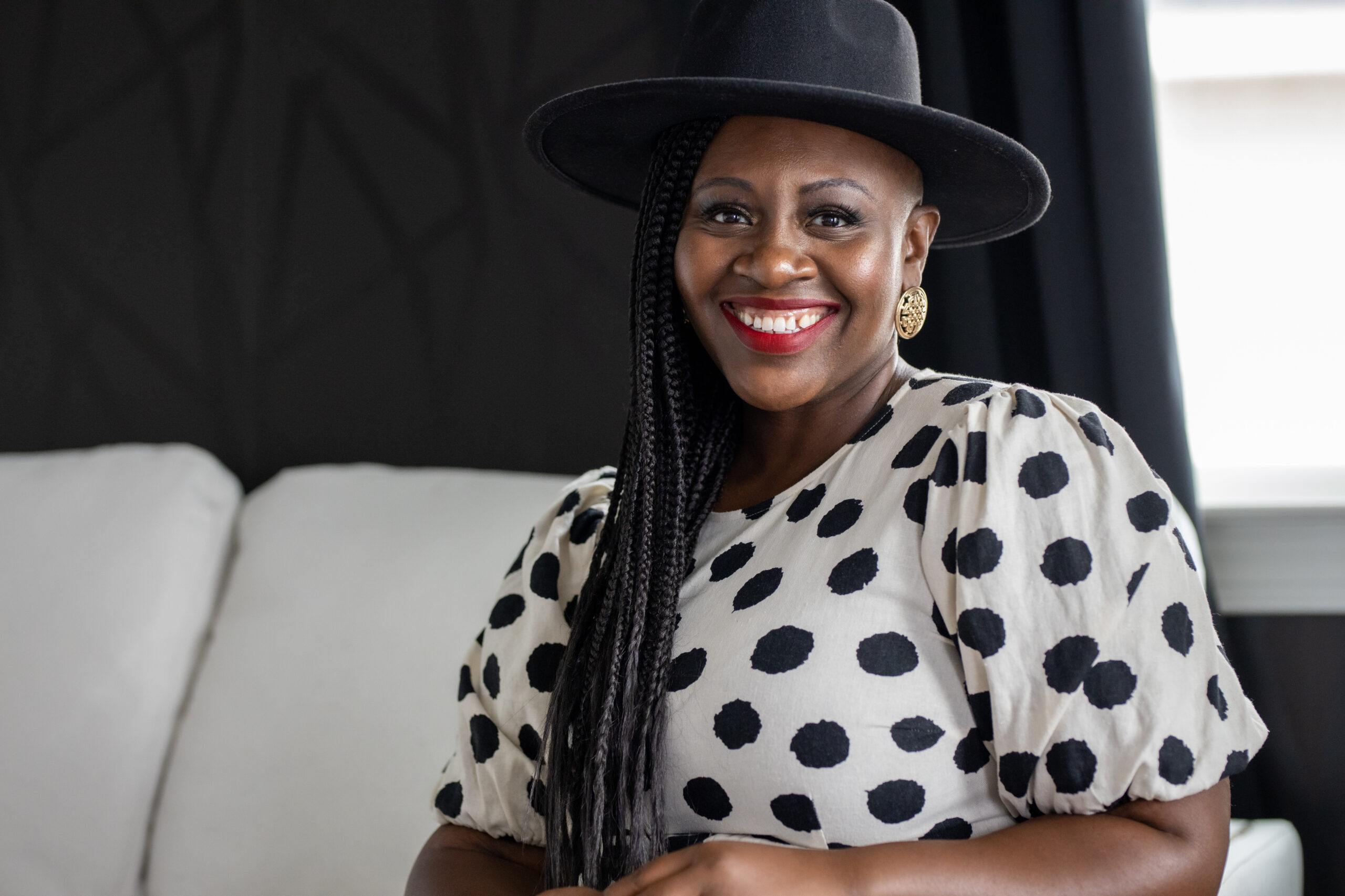 Pamela is a brown skin african american woman. She is sitting on a white sofa. She has on a white dress with black polka dots. She has on a black hat and is wearing red lipstick.